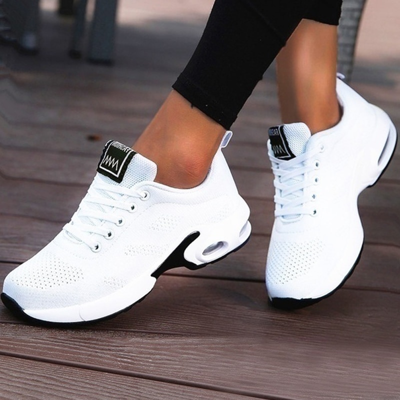 Scarpe da ginnastica bianche con suola bianca, etichetta nera sul davanti e lacci bianchi con dettagli neri sulla suola sui piedi incrociati di una donna che indossa leggings neri su un pavimento di parquet marrone.